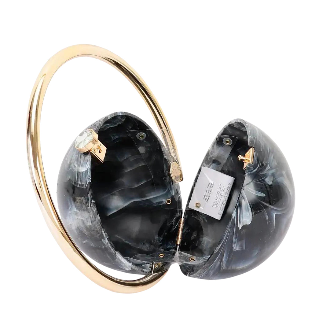 A split-hem clutch bag made of black and gray marbled resin, held together by a wide, polished gold-tone metal arch. A small gold clasp and a white product tag are visible on the inside of the halves.